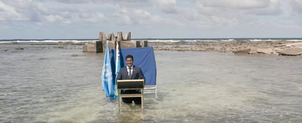 Photo of Hon Minister Simon Kofe standing in ocean with a podium